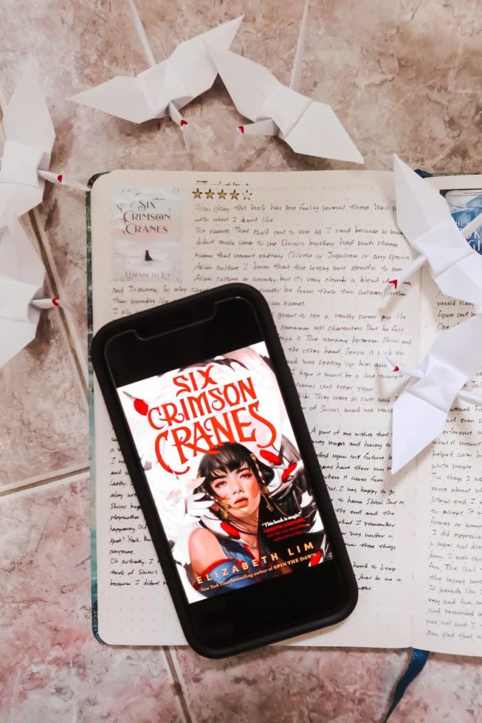 a phone with the cover of six crimson cranes lays on top of an open reading journal. there are six paper cranes with red crowns surrounding the phone and journal