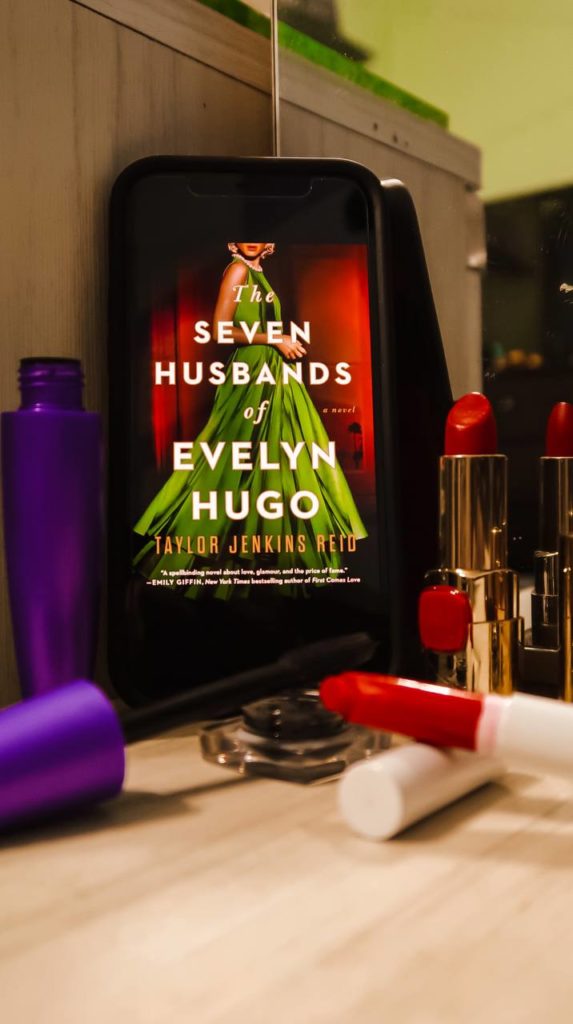 a phone with the cover of "the seven husbands of evelyn hugo" propped up. around it are different makeup products
