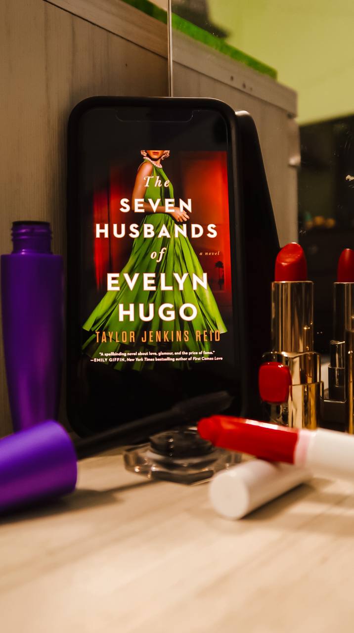 a phone with the cover of "the seven husbands of evelyn hugo" propped up. around it are different makeup products