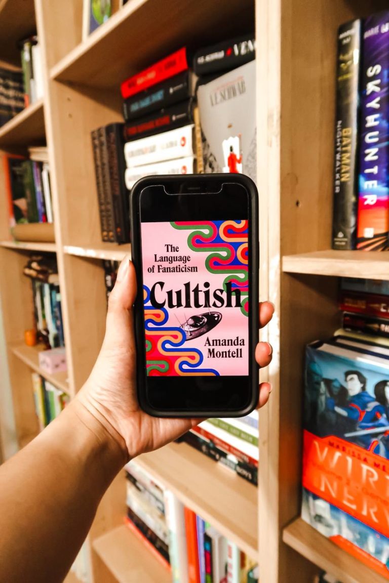 a phone featuring the cover of cultish by amanda montell held against a wall of bookshelves