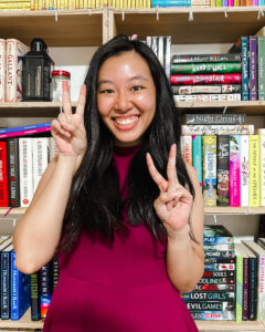 Charmaine standing in front of a bookshelf, smiling and in a red dress. She has two fingers held up on each hand